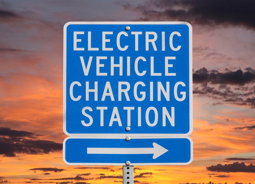 Electric vehicle charging station sign isolated with sunset sky.